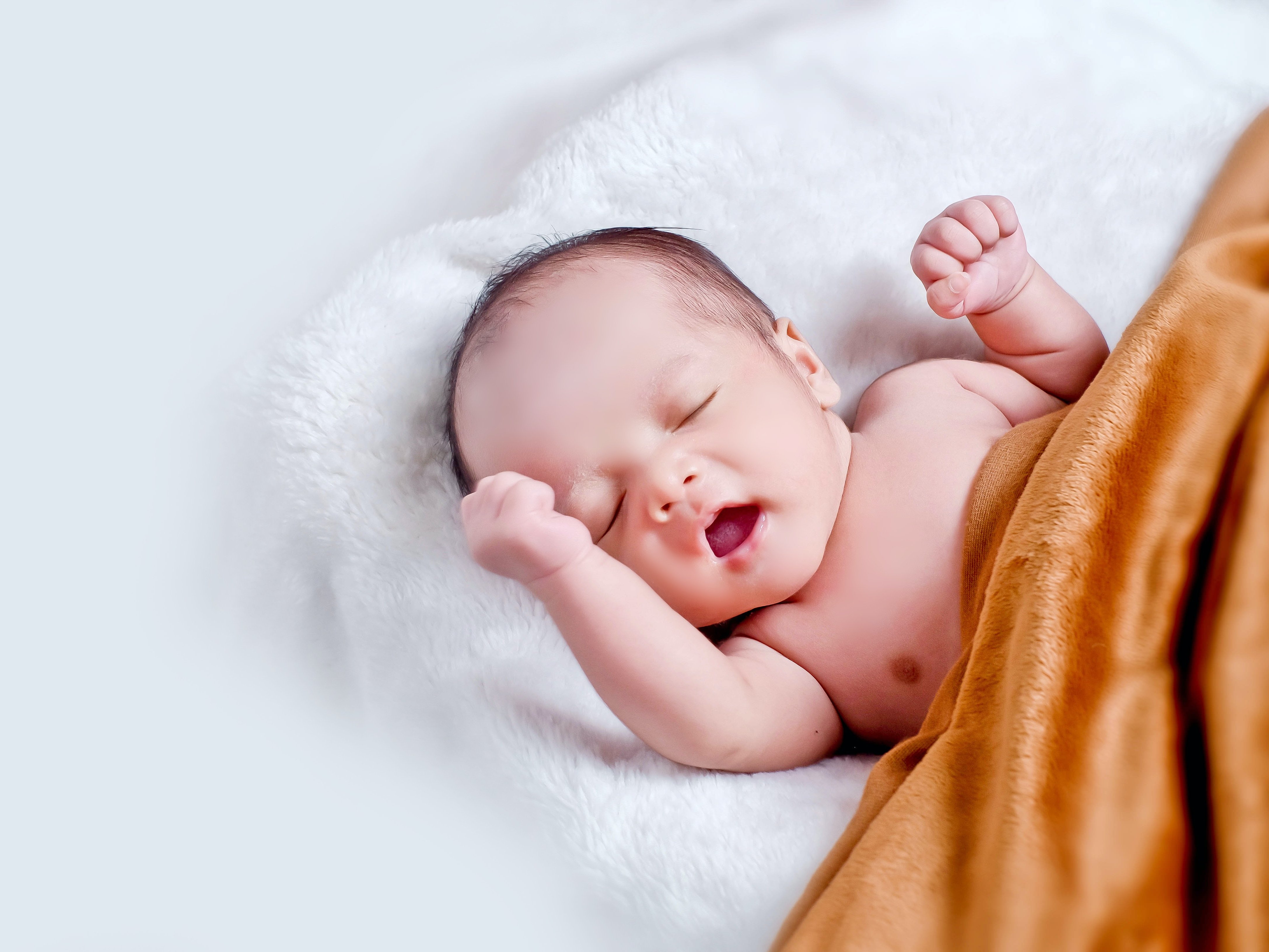 A baby lies with its back on a white blanket and is covered with a brown blanket. 